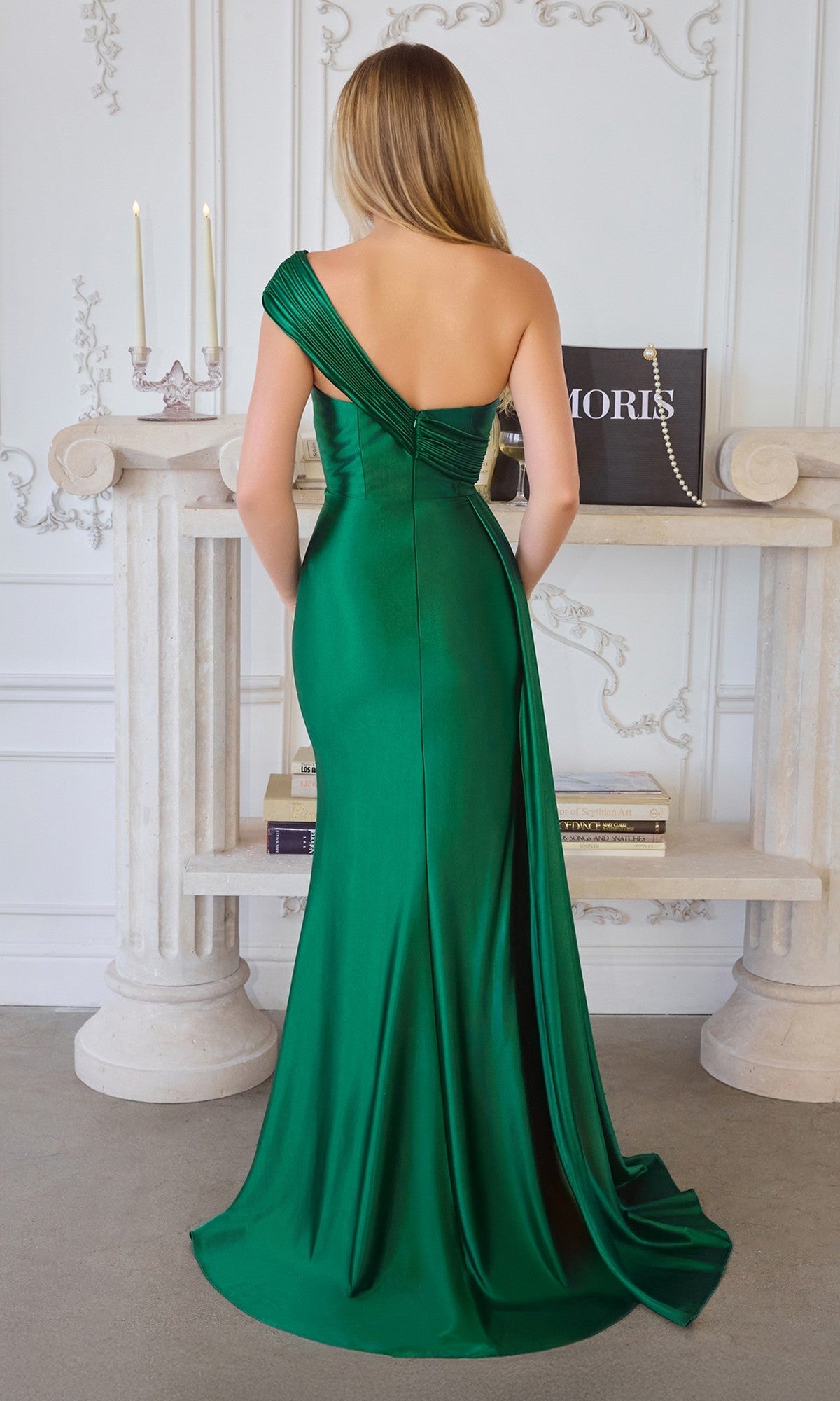 A woman in the Amoris T817 One-Shoulder Long Chiffon Prom Dress in green stands with her back to the camera in an elegant room decorated with candles, books, and ornate wall details.